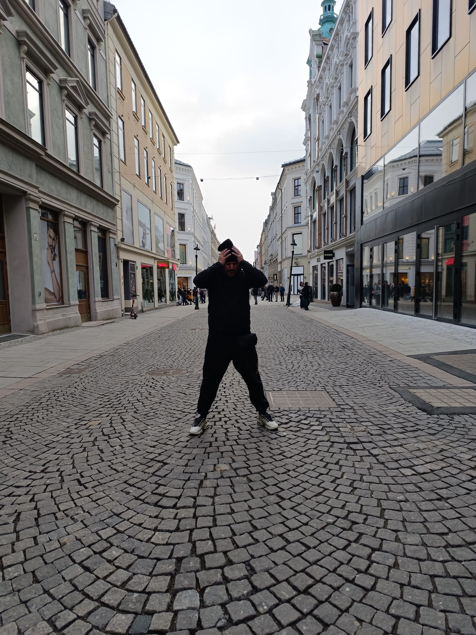 Enrique de pie en una calle urbana europea, vestido de oscuro, ajustándose el gorro, encuadre de cuerpo entero entre fachadas