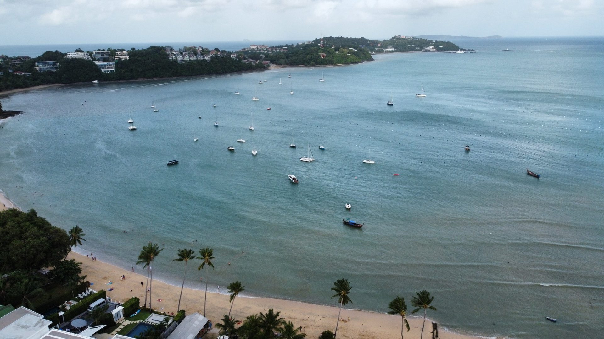Vista de dron de la bahía de Cape Panwa con decenas de veleros fondeados, playa con palmeras y el cabo al fondo