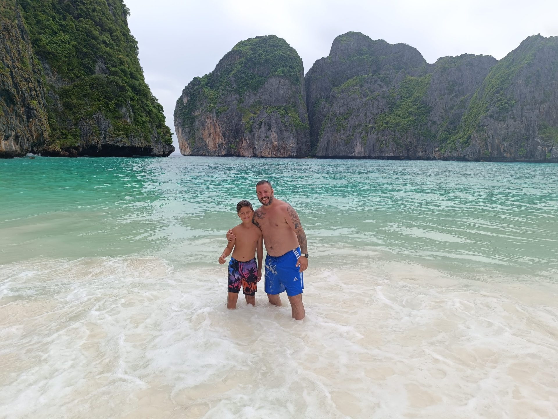 FUNESMA79 y su hijo en la orilla de una playa de arena blanca de Phi Phi, con agua turquesa y paredes de caliza detrás
